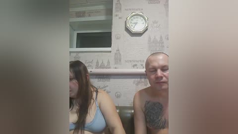 A video captures two people in a dimly-lit room with a clock and newspaper-themed wallpaper. A woman with long brown hair and a blue bra sits, while a bald man with a tattooed chest stands behind her.