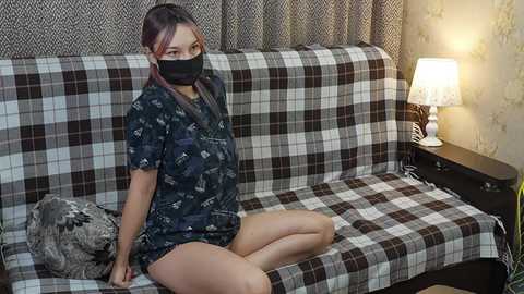 Video of an Asian woman in a black face mask, dark patterned pajamas, sitting on a plaid couch in a cozy room with a warm-lit table lamp.