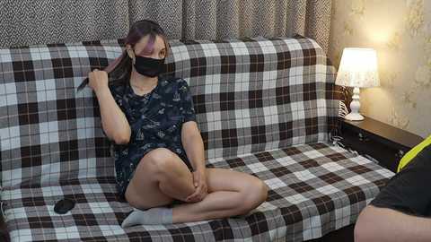 Video of a young Asian woman with light skin, wearing a face mask and black pajamas, sitting on a plaid couch, brushing her hair, in a cozy room with a lamp and patterned wallpaper.