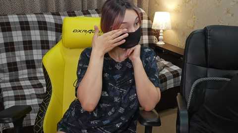 Video of a woman in a dark shirt, wearing a black face mask, sitting on a yellow gaming chair, in a cluttered room with a lamp and patterned wallpaper.