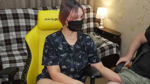 Video of a woman with short, dyed pink hair, wearing a black face mask, black patterned t-shirt, and sitting in a yellow gaming chair, next to a man in a green shirt, in a cozy, plaid-covered room with a lit lamp.
