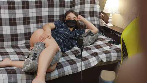 Video of a young woman with fair skin and long brown hair, wearing a black face mask, navy blue shirt, and gray shorts, lying on a plaid-patterned couch, recording herself with a black microphone.