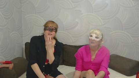 Video of two women, one in black, the other in pink, wearing white lace masks, sitting on a brown couch in a room with patterned wallpaper.