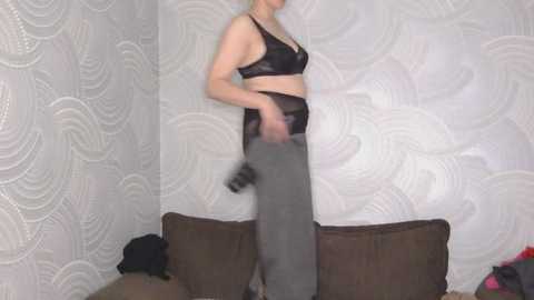 Video of a fair-skinned woman with blonde hair in black lingerie, grey pants, and a black handbag, standing in a room with patterned wallpaper and a brown couch.