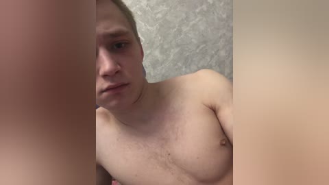 A video of a shirtless, fair-skinned man with short hair, looking intently at something off-camera. Background shows a textured gray wall.