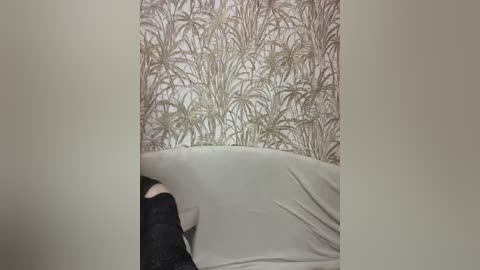 A video of a woman with light skin and dark hair, wearing a black sleeveless top, lying on a beige bed with a patterned wallpaper featuring palm trees in shades of brown and green.