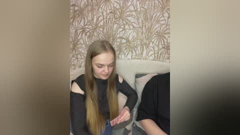 Video of a young woman with long, straight, blonde hair wearing a black top, adjusting a red and white striped scarf. Background shows a beige headboard and wallpaper with a palm tree pattern.