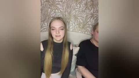 Video of a young woman with long, straight blonde hair, fair skin, and a black turtleneck sweater. She sits next to a man with short, light brown hair and a black t-shirt, both against a beige wallpaper with a leaf pattern.