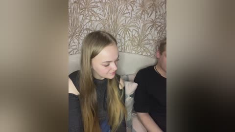 Video of a young Caucasian woman with long blonde hair, wearing a black off-shoulder top, sitting beside a man on a beige sofa, against a wallpaper with brown and gold palm leaf pattern.