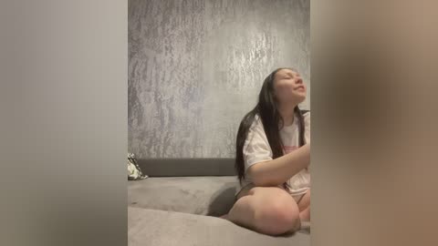 Video of a young Asian woman with long dark hair, wearing a white floral dress, sitting on a gray couch against a textured gray wall, eyes closed, serene expression.