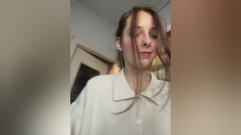 Video of a young woman with light skin and brown hair, wearing a beige cardigan, smiling softly, standing in a dimly lit, indoor room with a wooden door and blurred background.