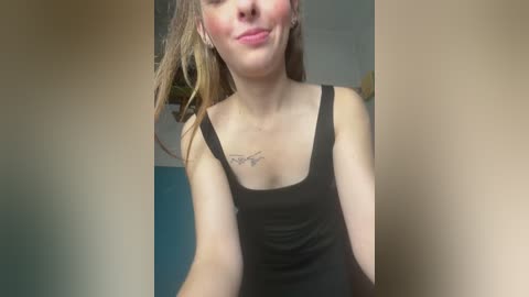 A video of a smiling young woman with fair skin and light brown hair, wearing a black tank top, standing indoors against a blue wall. She has a small tattoo on her chest.