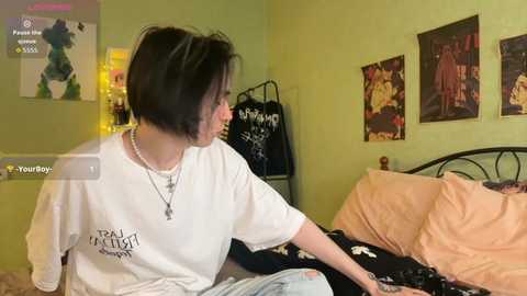 Video of a young woman with short black hair, wearing a white T-shirt and jeans, sitting on a bed with peach-colored sheets and dark floral bedspread in a dimly lit room.