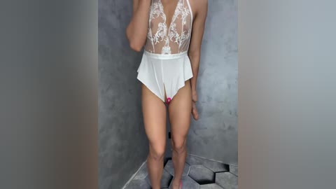 Video of a slender woman with medium skin tone wearing a sheer, lace bodysuit with a plunging neckline and high-cut legs, posing in a minimalist, concrete-walled shower with hexagonal tiles.