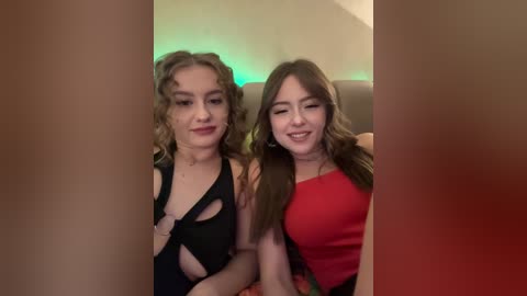 Video of two young women with curly blonde and long brown hair, smiling, seated in dimly lit room with green lighting, wearing black and red dresses.