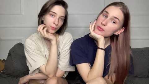 Video of two young women, one with short brown hair, the other with long brown hair, both with fair skin and small breasts, resting their chins on their hands, sitting on a gray couch against a white wall.