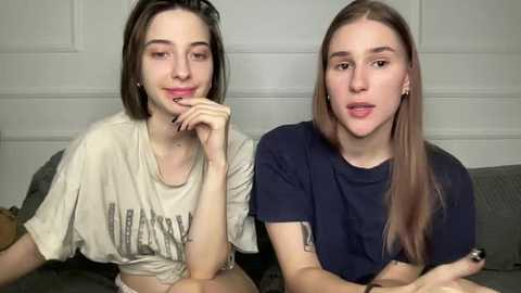 Video of two young women with straight, light brown hair, one in a white shirt, the other in a dark blue shirt, sitting side by side on a couch.