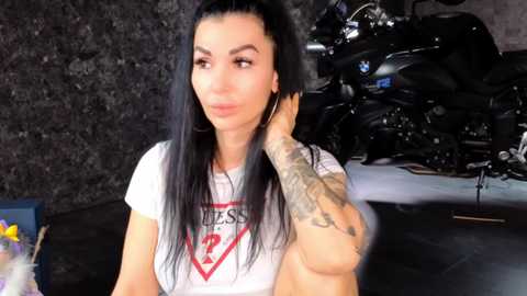 Video of a tattooed woman with long black hair, wearing a white t-shirt, sitting indoors next to a motorcycle.