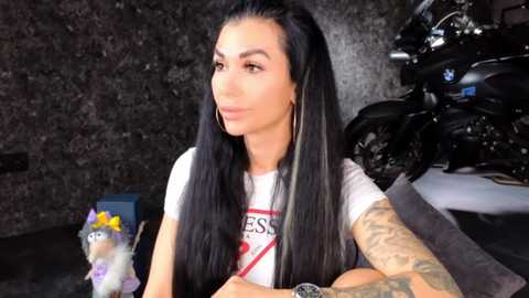 Video of a tattooed woman with long black hair, wearing a white t-shirt, seated in a dimly lit room with a motorcycle and a plush toy in the background.