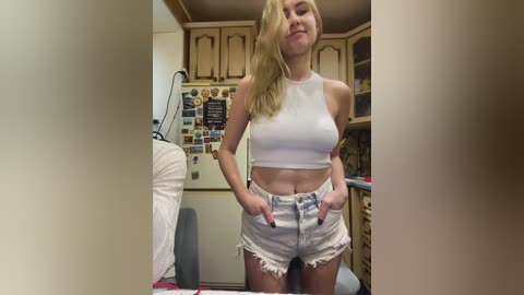 Video of a blonde woman in a white crop top and distressed denim shorts, standing in a cluttered, beige kitchen with wooden cabinets and various posters on the wall.