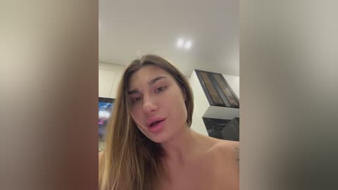 A video of a young woman with light skin, long brown hair, and bare shoulders, taken from a slight angle, in a modern, dimly lit room with a TV and abstract artwork in the background.