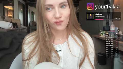 Video of a young Caucasian woman with long, straight, light brown hair and blue eyes, wearing a white sweater, making a pouty face in a modern, well-lit living room with a TV displaying \"Go Low\" by Yung Thug.
