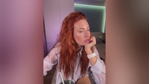 Video of a fair-skinned, red-haired woman with wet hair, wearing a white blouse, and a white bracelet, biting her finger, surrounded by blurred objects, in a modern room with purple and green lighting.