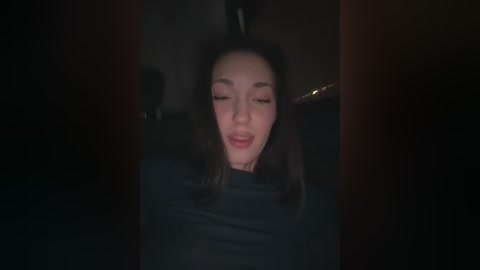 A dimly lit video of a young woman with fair skin and long, dark hair, wearing a black top, smiling slightly. Background is dark, with indistinct shapes, suggesting an indoor setting.