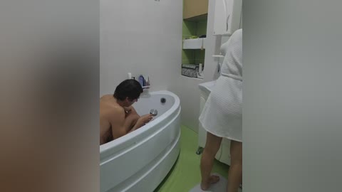 Video of a nude man in a white bathtub, washing his hair, in a modern bathroom with green tiles and a white robe hanging.
