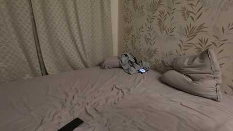 Video of a messy bedroom with a beige bedspread, a smartphone, and a pair of clothes on the bed, surrounded by patterned wallpaper and a closed curtain.