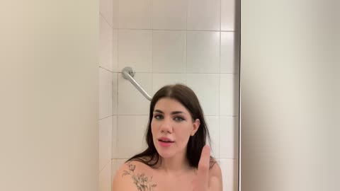 Video of a young woman with fair skin, long dark hair, and tattoos on her shoulders, standing in a white-tiled shower, wearing minimal makeup, with a slight smile.