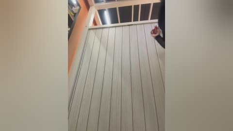 Video of a beige-painted wooden porch floor with vertical slats, featuring a partially visible person in a black shirt and red bag, standing on the right. The background includes a white railing and orange wall, with a framed picture visible on the left.