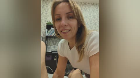 Video of a smiling, fair-skinned woman with shoulder-length blonde hair, wearing a white t-shirt, leaning over a table in a room with patterned wallpaper, bookshelves, and a green plant in the background.
