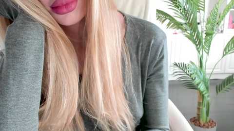 Video of a woman with long, platinum blonde hair, wearing a green V-neck top, leaning on her left elbow, lips slightly parted, in a bright, modern room with a green plant and white radiator in the background.