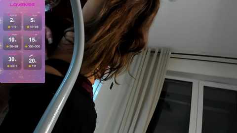 Video of a woman with long, wavy brown hair, wearing a black top, standing in front of a window with white curtains, showing a weather app on the left side.