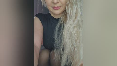 Video of a light-skinned woman with long, wavy blonde hair, wearing a black sleeveless top, sitting indoors against a blurred beige wall.