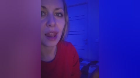 Video of a woman with blonde hair, light skin, and a red shirt, looking through a door with a blue light in the background.