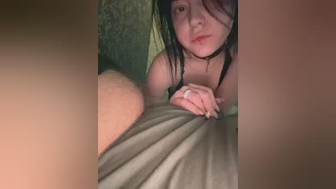 A close-up video of a young East Asian woman with long, wet black hair, lying on a bed, looking directly at the camera. She wears a black choker and a gray tank top.