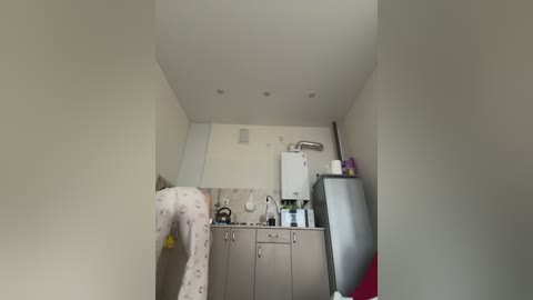 A video of a small, minimalistic kitchen with a white wall and beige cabinets. A person in white pajamas with a floral pattern is bent over a sink. A refrigerator and microwave are visible on the right.