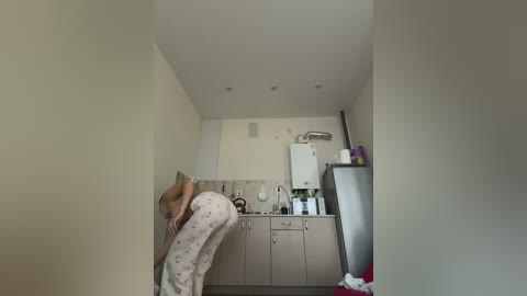 Video of a small, narrow kitchen with beige walls and cabinets. A person wearing light pajamas is bent over, possibly cleaning. White fridge, sink, and stove on the right.