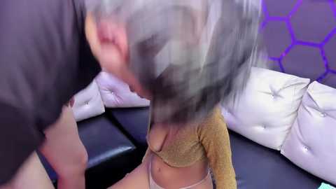 A blurry video of a woman with short silver hair, wearing a gold bralette, and a man with dark hair, both on a black couch.