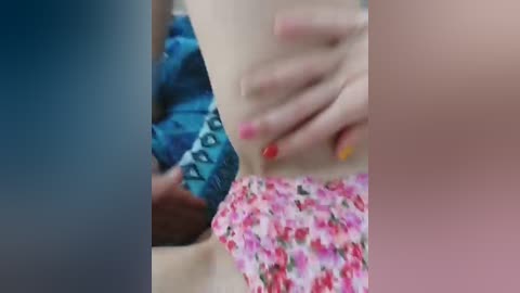 A close-up video of three hands with different nail polish colors: red, yellow, and pink, placed on a person's bare chest, partially covered by a floral-patterned fabric. The background is blurred, focusing on the intimate interaction.