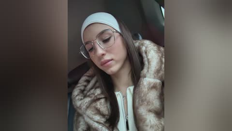 Video of a young woman with light skin, long straight brown hair, wearing clear glasses, a white headband, and a brown faux fur coat, seated indoors, looking down.