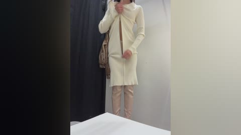 Video of a person standing in a fitting room, wearing a cream-colored, knee-length sweater dress and beige tights, holding a brown handbag. The background is a plain white wall with a black curtain.