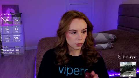 Video of a young woman with wavy brown hair, fair skin, and a black \"Viper\" sweatshirt, sitting on a bed in a dimly lit room with purple lighting.