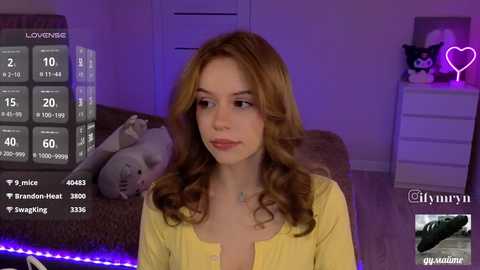 Video of a young woman with wavy brown hair and fair skin, wearing a yellow top, sitting on a couch in a dimly lit room with purple lighting, plush toys, and a heart-shaped neon sign.