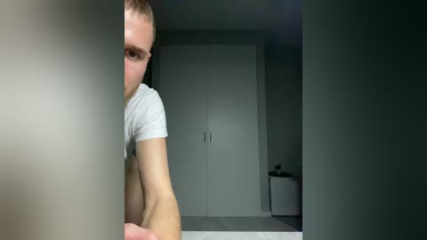 A video of a young man with short brown hair and a beard, wearing a white T-shirt, crouched in a dimly lit room with white cabinets and a small trash bin in the background.