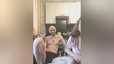 Video of a bald, shirtless man with a tattooed chest, sitting in a kitchen, holding a black object, with a woman partially visible in the foreground.