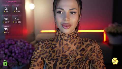 Video of a young woman with light brown skin, dark hair, and dark eyes, wearing a leopard print top, set in a dimly lit room with purple flowers and a blurred background.