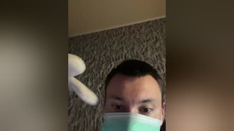 A video shows a man wearing a blue surgical mask, looking up, with a white stuffed bunny on his head. The background features a beige carpet and a beige wall corner.